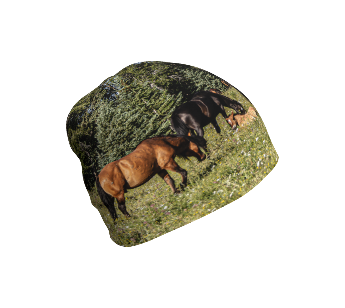 This is a beanie featuring a photograph of wild foals lying in wildflowers in the summer, with a herd of wild horses grazing in a field surrounded by trees. Color tones include greens, browns, and black.