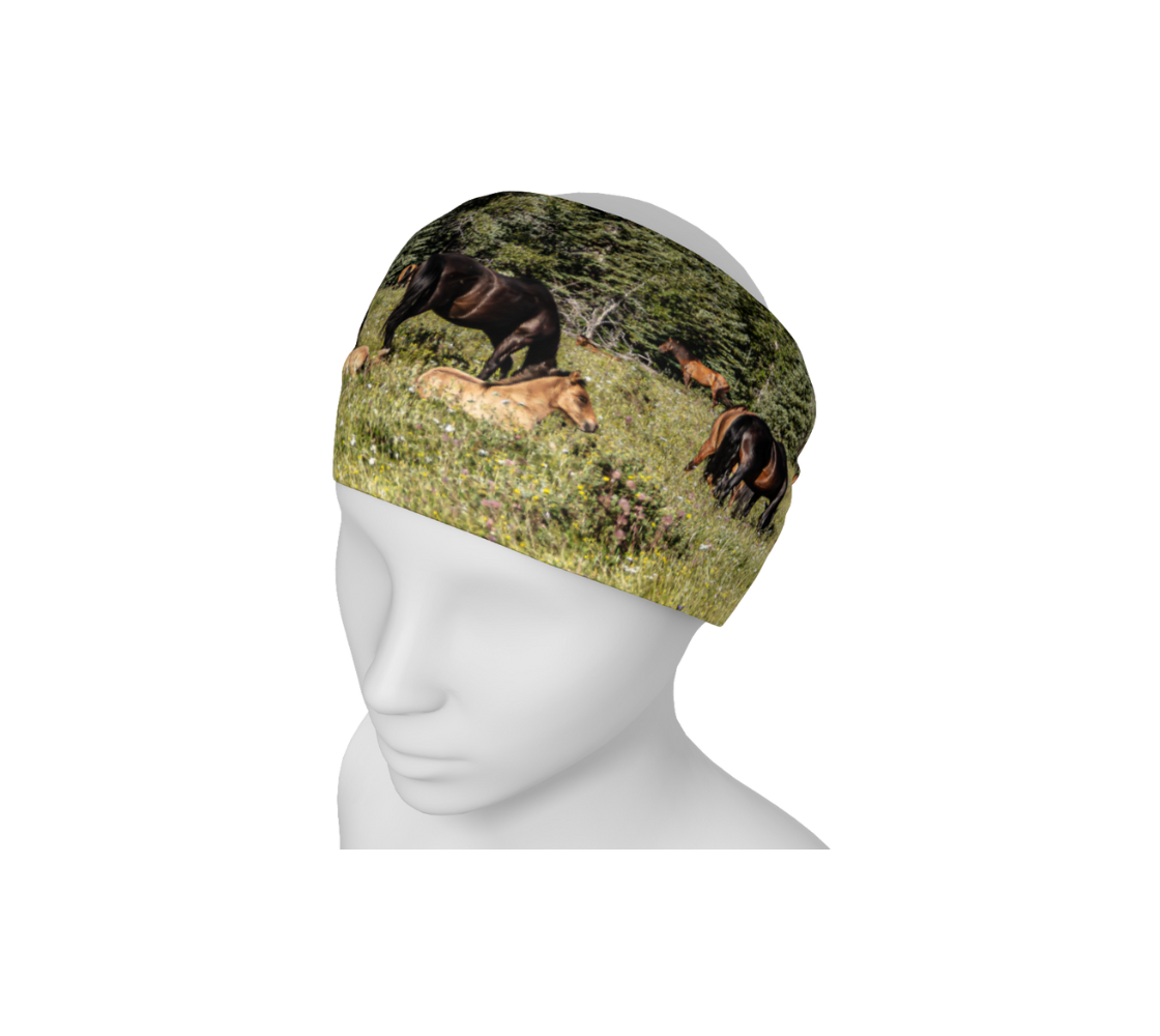 This is a head and neck band of a herd of wild horses on a mannequin head worn as a headband. It features a photograph of wild foals lying in wildflowers in the summer, with a herd of wild horses grazing in a field surrounded by trees with a blue sky. Color tones include greens, browns, blue and black.