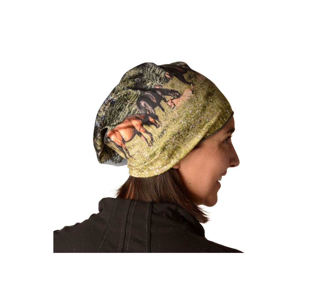 This is side profile of a woman wearing a photo of a herd of wild horses on a head and neck band as a kerchief. The image feaures wild foals lying in wildflowers in the summer, with a herd of wild horses grazing in a field surrounded by trees, with a blue sky. Color tones include greens, browns, and black.