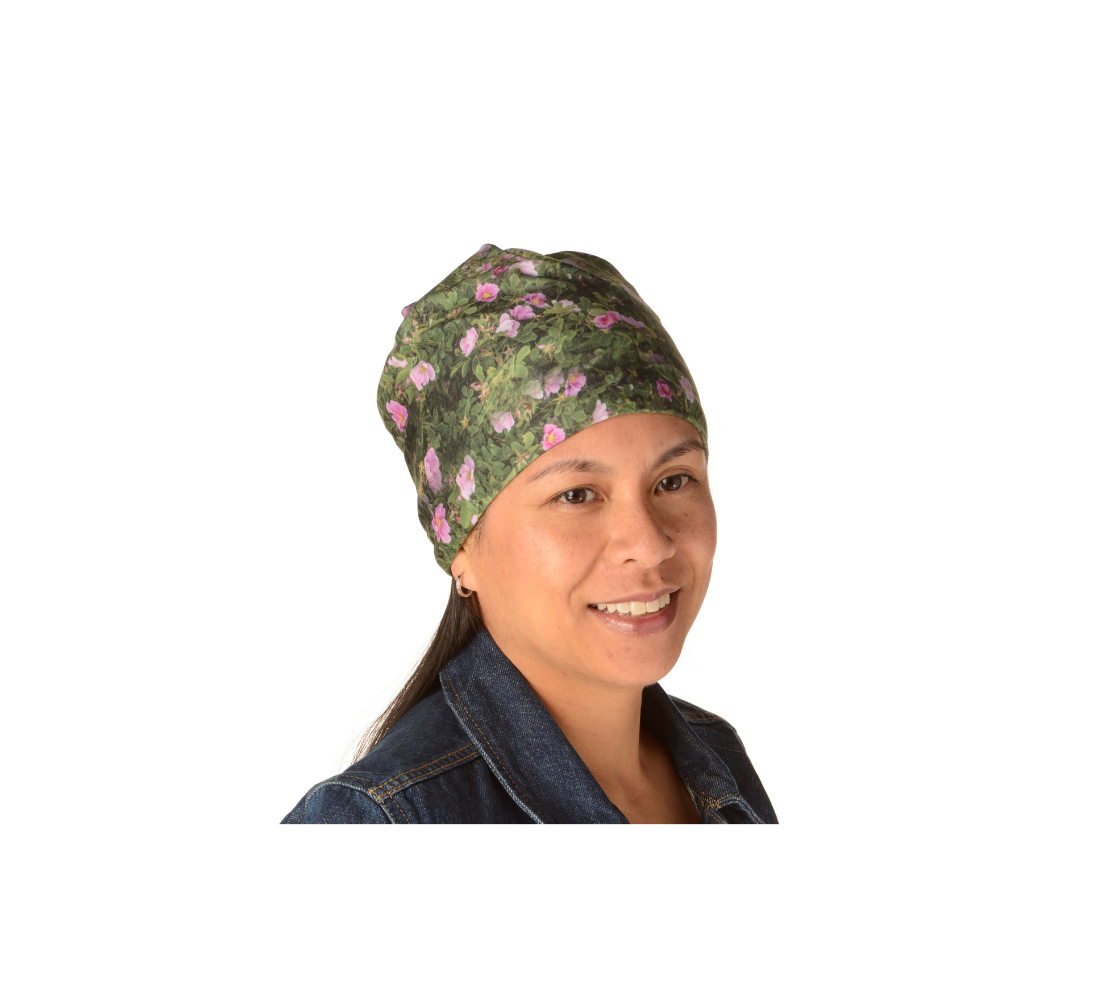 This is a front portrait of a lady wearing a photo of wild roses on a beanie. The beanie is a collage of green wild rose bushes with pink flowers and buds througout that have yellow centers.