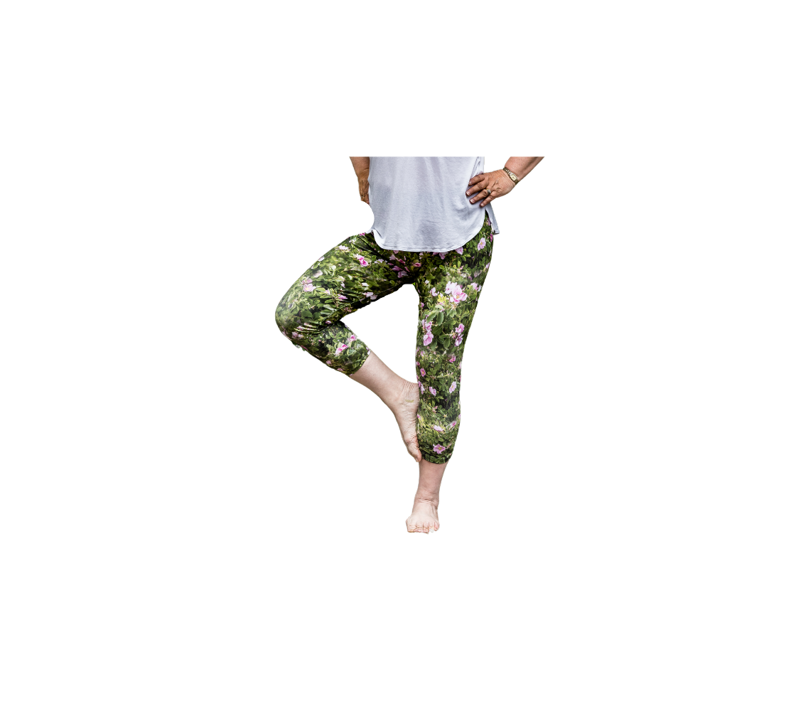 This is a front profile of a lady wearing a pair of 3/4 length wild rose capri leggings. The print is a photo collage of green wild rose bushes with pink flowers and buds througout that have yellow centers.