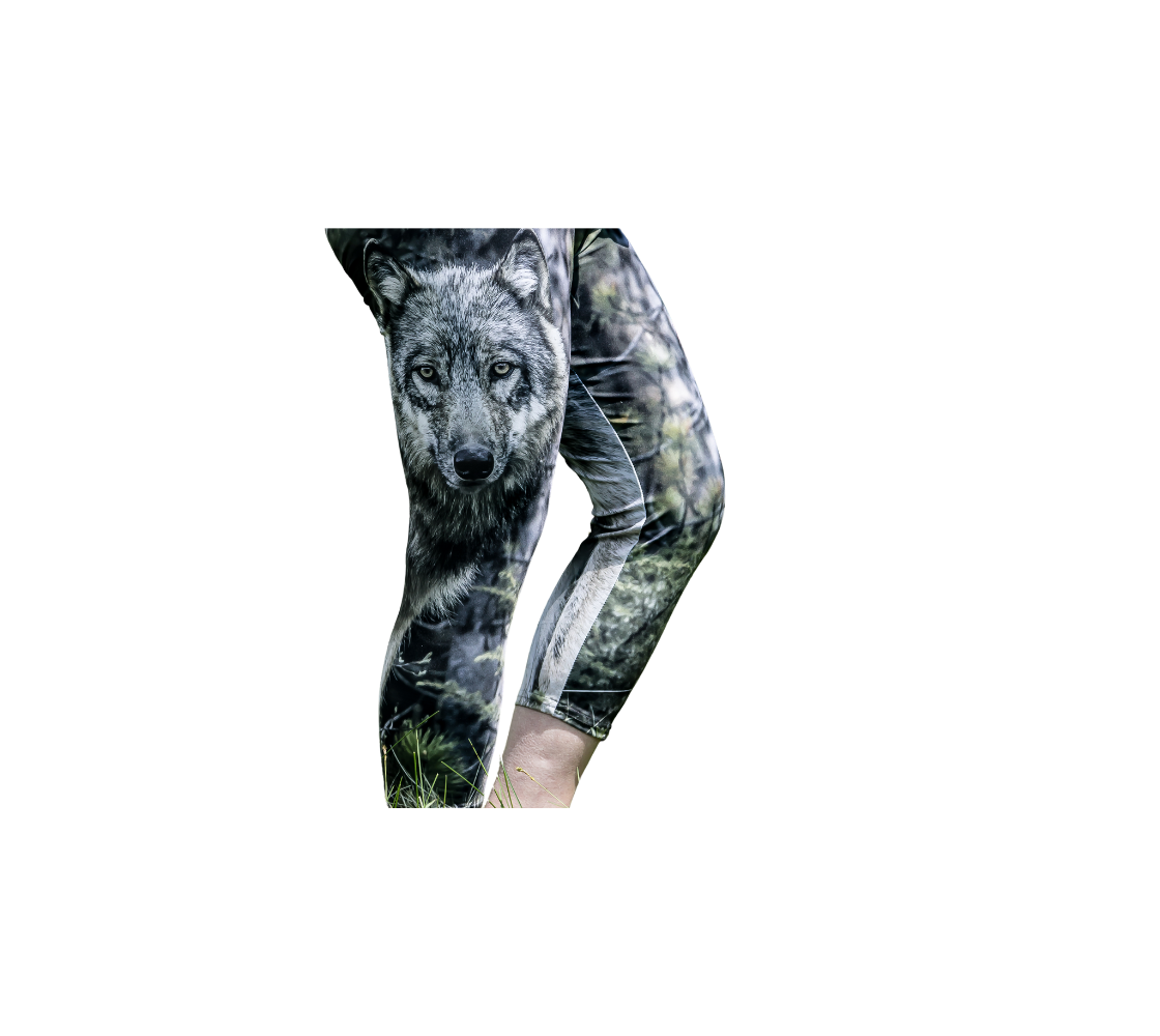 This is a side profile of a person wearing a pair of 3/4 length capri leggings featuring a photo of a wolf. It has the wolf head image on the side of the leg. The background is of evergreen trees and grey bushes, while the wolf head is full and fluffy in various tones of grey and white with beautiufl yellow eyes looking directly at you.
