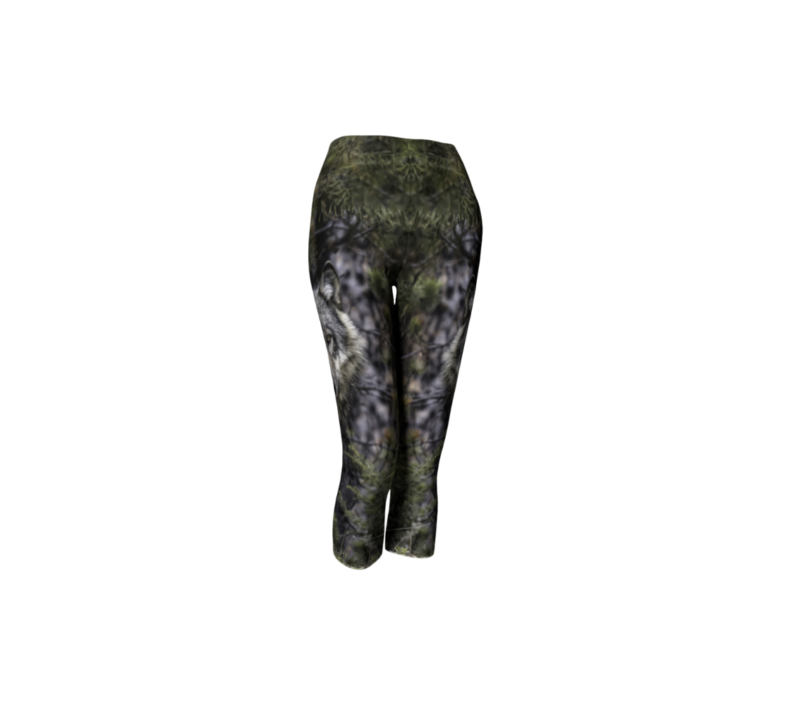 This is a front profile of a photo of a wolf head on a pair of 3/4 length capri leggings. It has the wolf head image on the side of each leg. The background is of evergreen trees and grey bushes, while the wolf head is full and fluffy in various tones of grey and white with beautiufl yellow eyes looking directly at you.