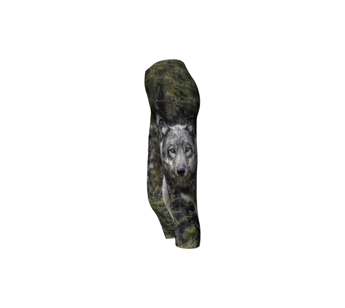 This is a side profile of a photo of a wolf head on a pair of 3/4 length capri leggings. It has the wolf head image on the side of the leg. The background is of evergreen trees and grey bushes, while the wolf head is full and fluffy in various tones of grey and white with beautiufl yellow eyes looking directly at you.