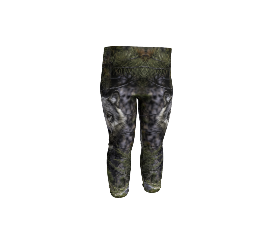 This is a front profile of a photo of a wolf head on a pair of infant leggings. It has one wolf head image on the side of each leg. The background is of evergreen trees and grey bushes, while the wolf head is full and fluffy in various tones of grey and white with beautiufl yellow eyes looking directly at you.
