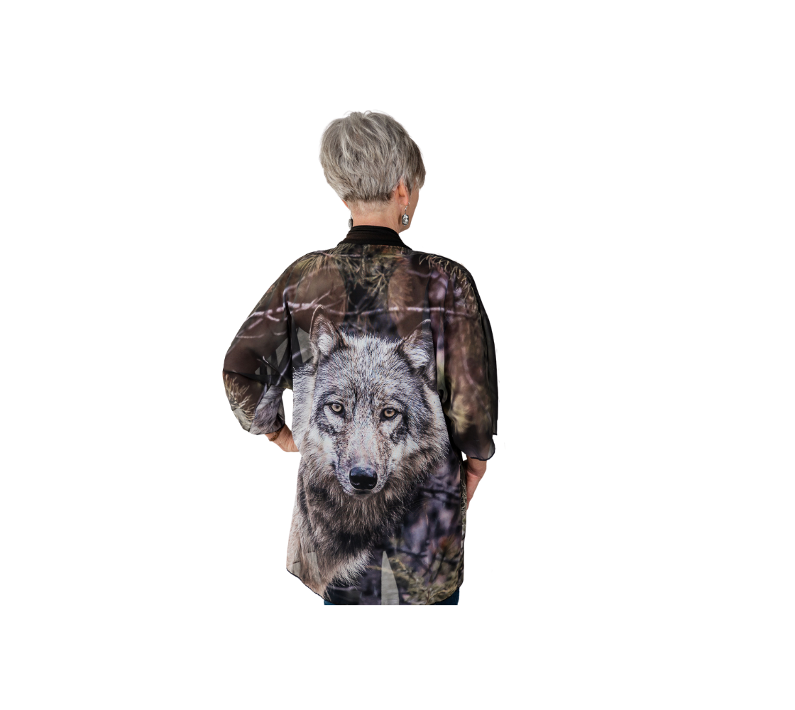 This backview is of a lady wearing a semi-sheer poly chiffon kimono robe  featuring a photo of a wolf. Background is green and grey with pine tree branches with a grey wolf head shot. Kimono has black lightweight bamboo trim.