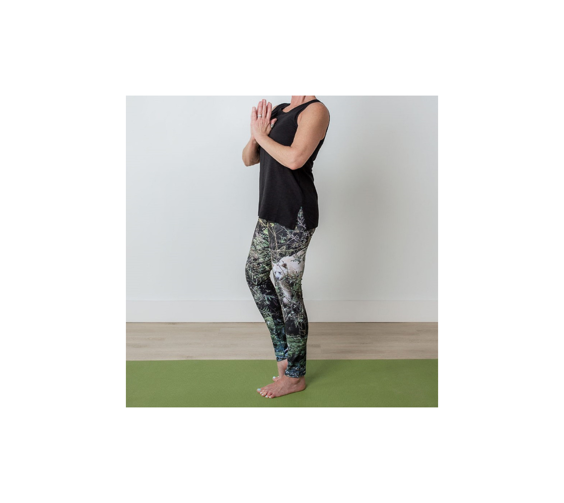 This is a side profile of a lady in a pair of yoga leggings featuring a photo of a spirit bear on each leg, with a 4.5 inch wide waistband. The background is green bushes and brown branches of the Great Bear Rainforest. The white spirit bear is stepping down out of the forest and is making her way to the stream below.