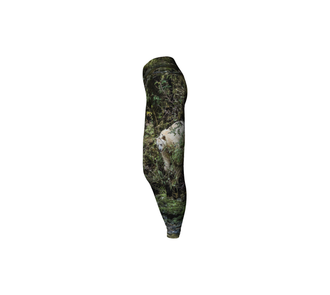 This is a side profile of a pair of yoga leggings featuring a photo of a spirit bear on each leg, with a 4.5 inch wide waistband. The background is green bushes and brown branches of the Great Bear Rainforest. The white spirit bear is stepping down out of the forest and is making her way to the stream below.