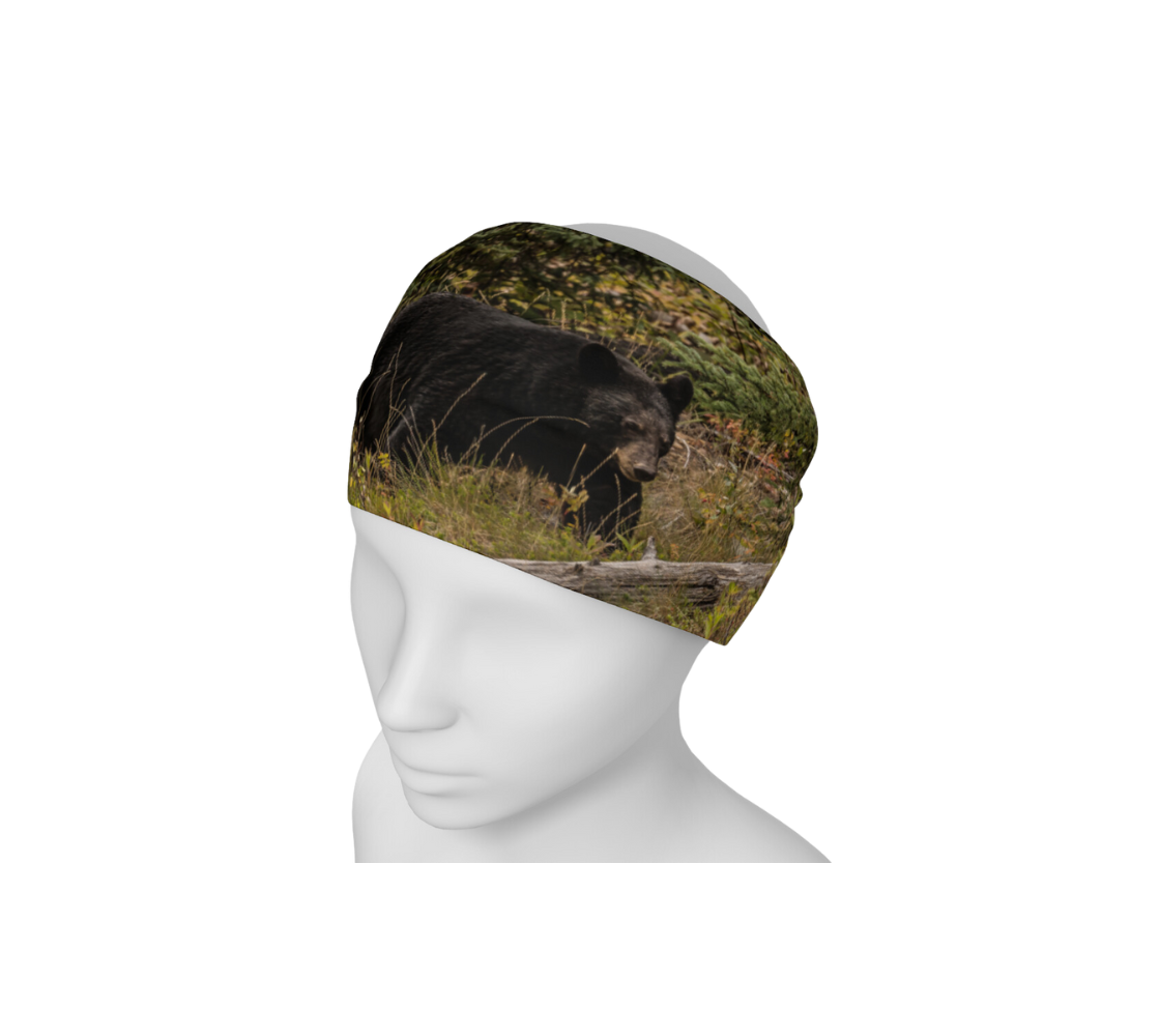 This is a photograph of a black bear on a head and neck band, worn as a headband on a mannequin. The large black bear is walking along a hill covered in green grasses and bushes with a old grey log lying in front of him. 