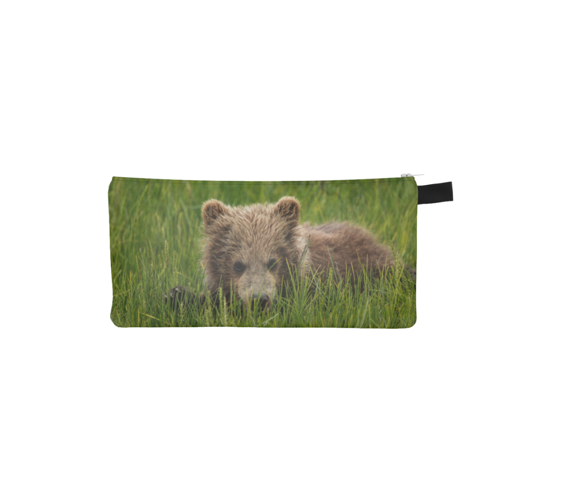 This is a photo on a zippered pouch of little brown bear cub hiding behind a blade of grass in the tall green grass. The background is bright green grass with a little light brown colored bear cub.