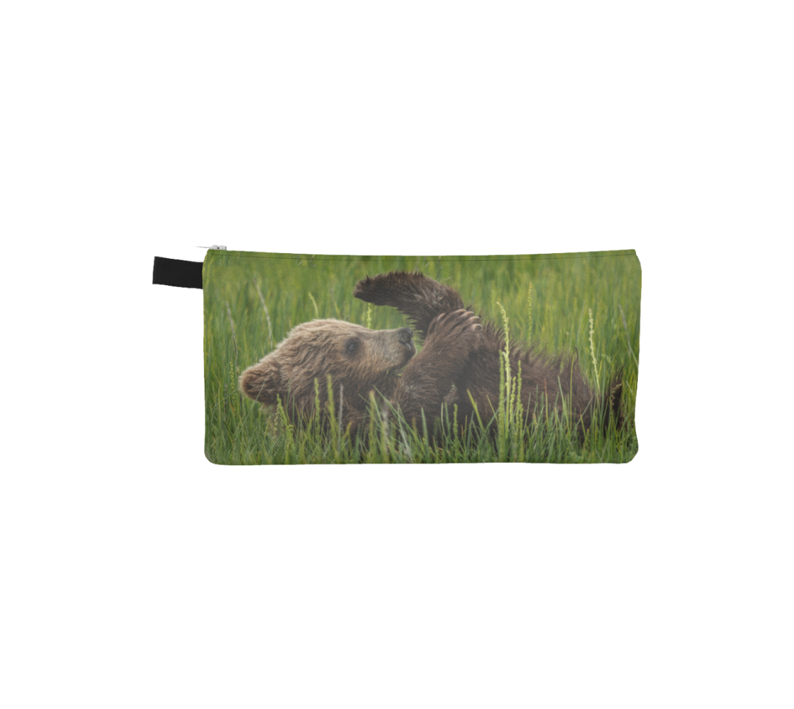 This is a photo on a zippered pouch of little brown bear cub lying back and playing with his feet in the tall green grass. The background is bright green grass with a little light brown colored bear cub.