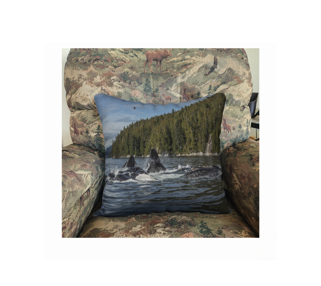 A 18 x 18 inch plush velveteen cushion cover features a pod of hump-back whale breaching surface of ocean and the background is green and blue. This image is of a cushion cover on a pillow form in a chair.