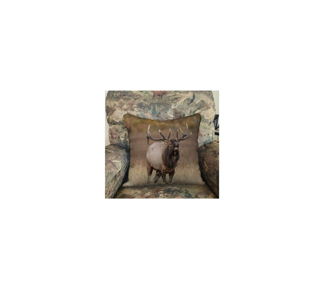 A 18 x 18 inch plush velveteen cushion cover featuring a photo of a bull elk bugling. The background is blurred yellow dead grass. The dark brown and tan colored elk with its large antlers is raising its head as it bugles out its mating call.. This image is of a cushion cover on a pillow form in a chair.