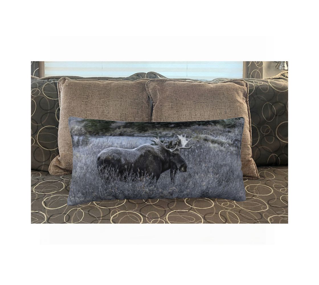 A 12 x 24 inch plush velveteen cushion cover features a photo of a bull moose covered in frost. This image shows a big dark brown bull moose standing in the frosty grey willows surrounded by evergreen trees. He is covered with a blanket of white frost on his back, shoulders and head.  This image is of a cushion cover on a pillow form on a bed.