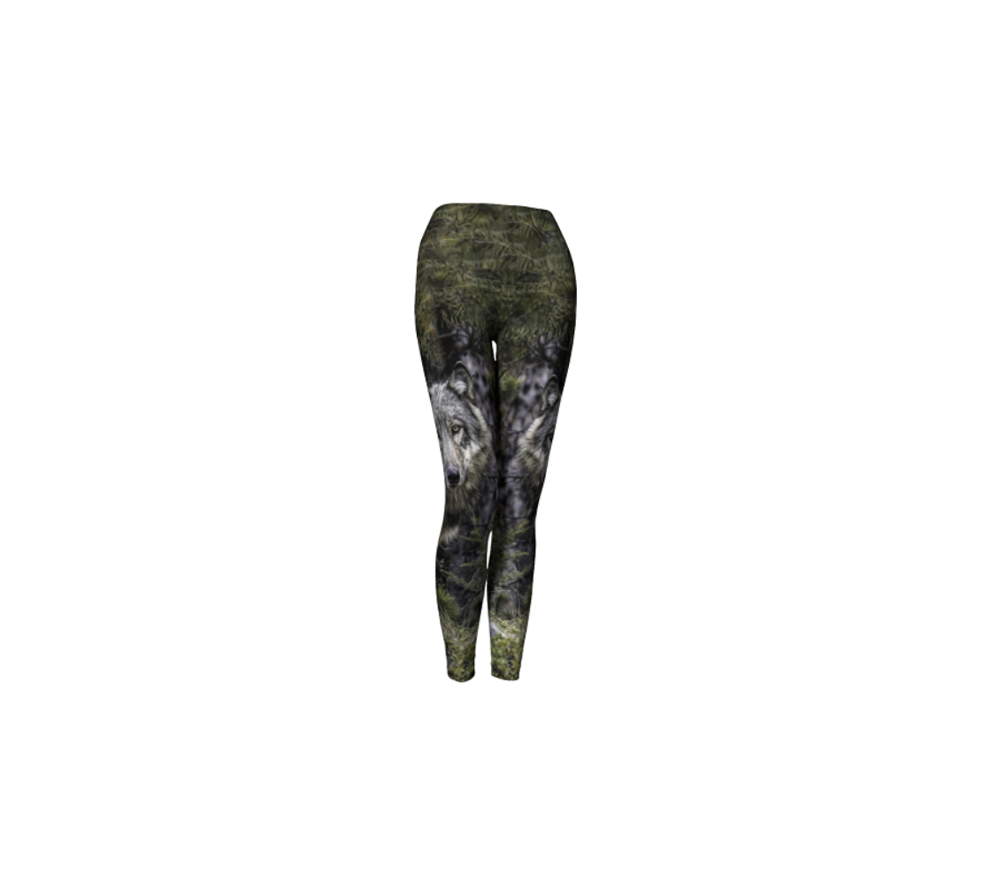 This is a side profile of a pair of yoga leggings with a wolf image on the side of each leg, with a 4.5 inch wide waistband. The background is of evergreen trees and grey bushes, while the wolf head is full and fluffy in various tones of grey and white with beautiufl yellow eyes looking directly at you.