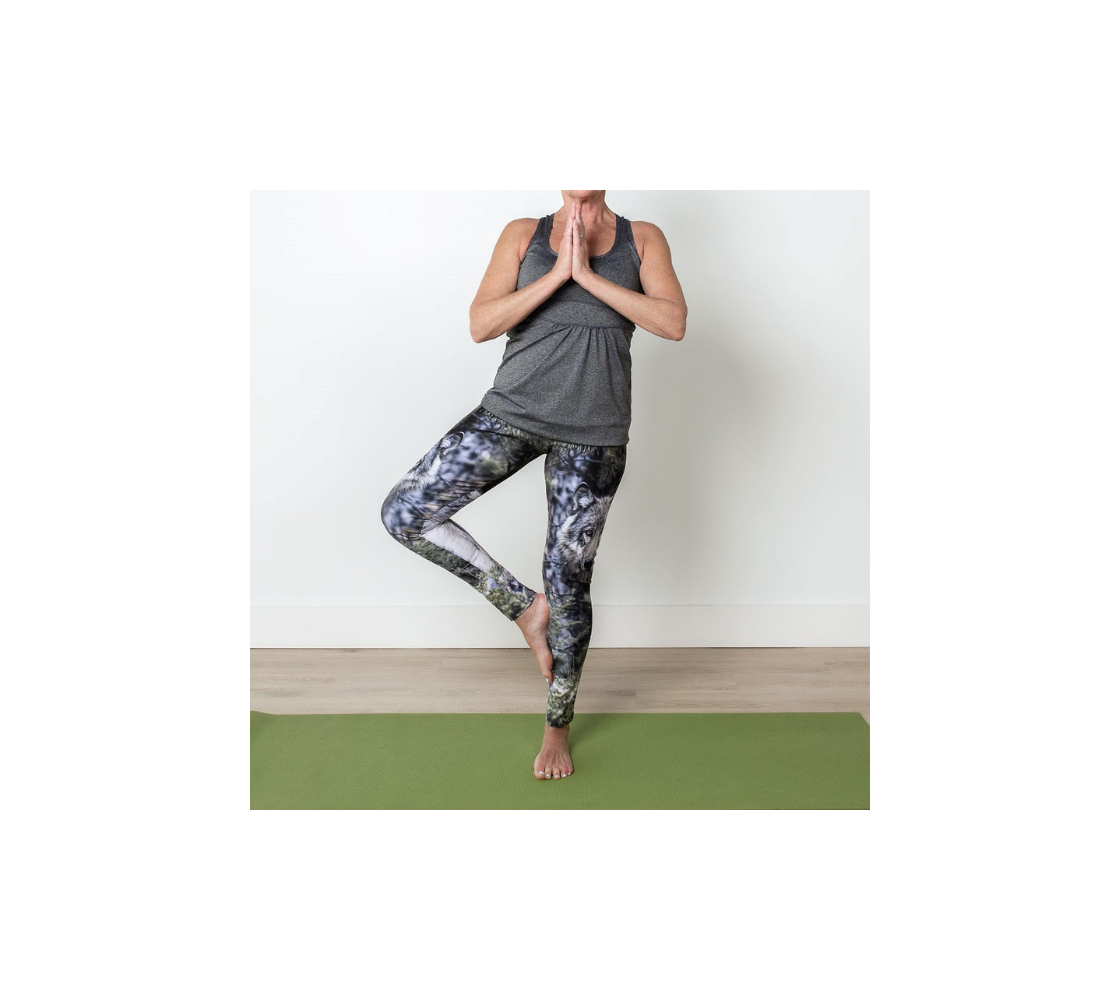 This is a front profile of a lady doing a tree yoga pose in a pair of yoga leggings with a wolf image on the side of each leg. The background is of evergreen trees and grey bushes, while the wolf head is full and fluffy in various tones of grey and white with beautiufl yellow eyes looking directly at you. 