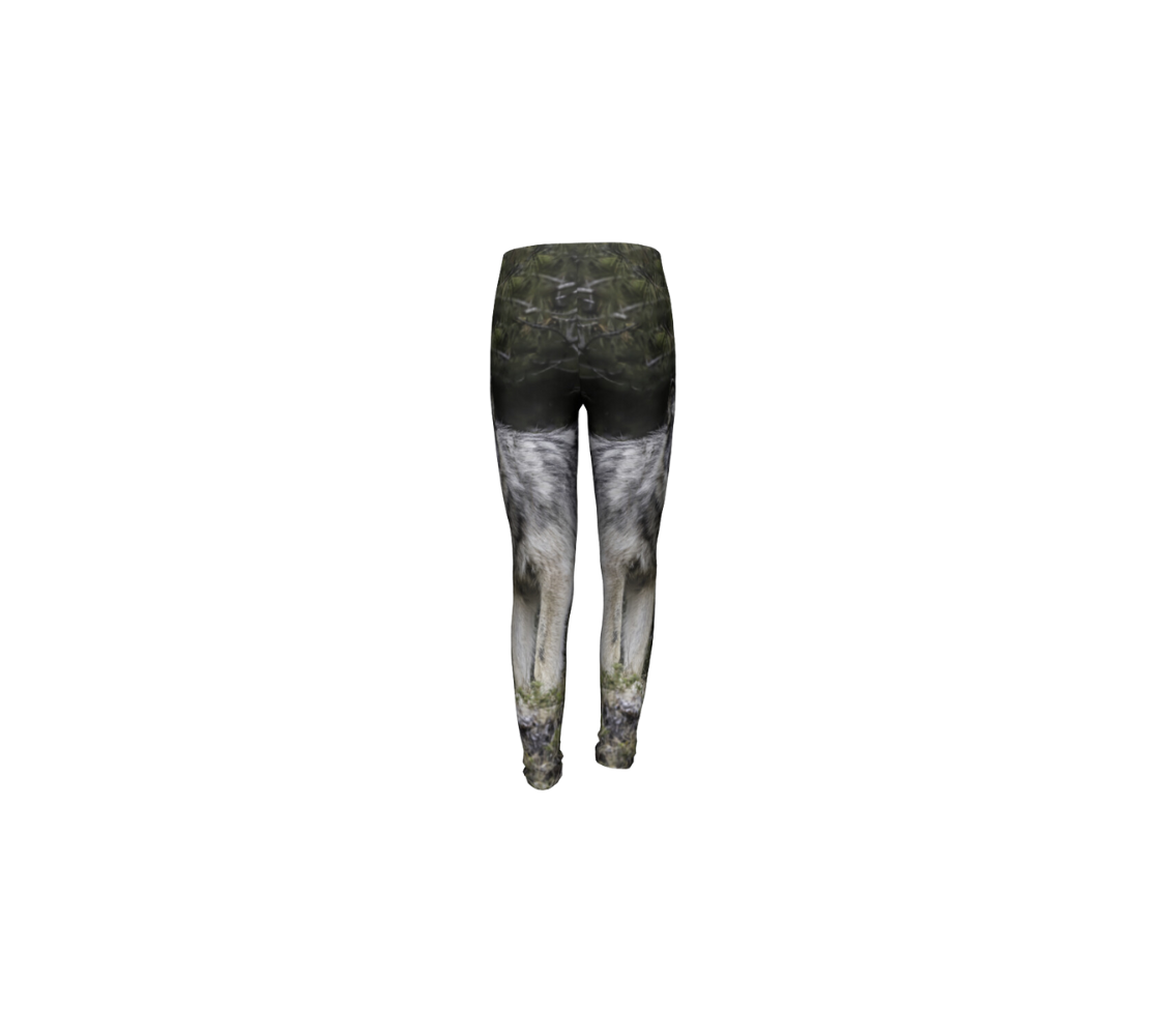 This is a rear profile of a wolf on a pair of kids leggings with a wolf image on the side of each leg. The background is of evergreen trees and grey bushes, while the wolf head is full and fluffy in various tones of grey and white with beautiufl yellow eyes looking directly at you.