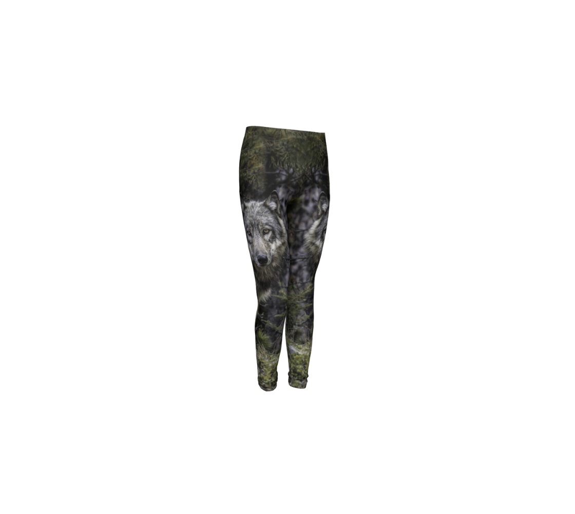 This is a front profile of a wolf on a pair of kids leggings with a wolf image on the side of each leg. The background is of evergreen trees and grey bushes, while the wolf head is full and fluffy in various tones of grey and white with beautiufl yellow eyes looking directly at you.