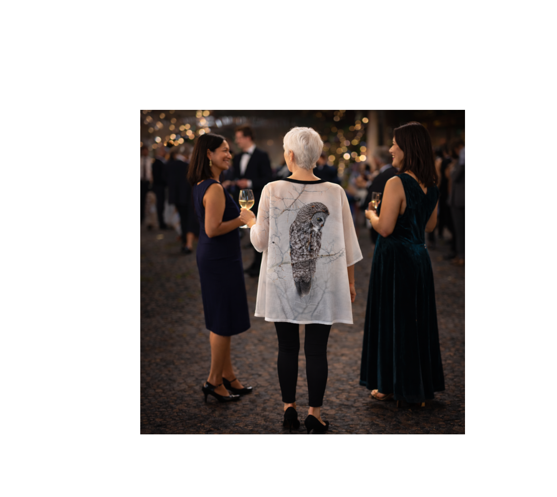 Woman in a kimono cover up with an image of a  great gray owl design with a white background standing among other people at an event.