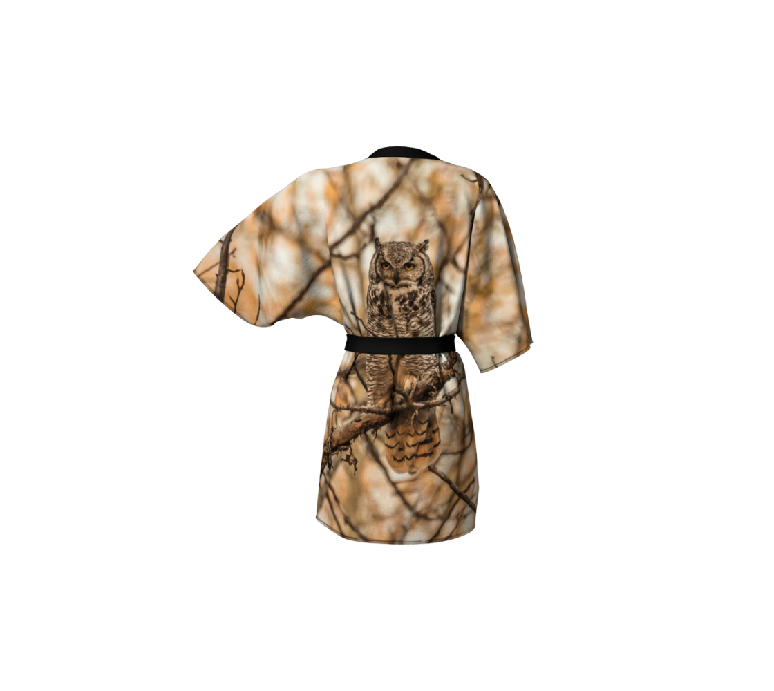 This backview is of a semi-sheer poly chiffon kimono robe featuring a photo of an owl. Background is of orange, brown and white blurred tree branches, with a great horned owl on a tree branch. Kimono has black trim and a black lightweight bamboo belt for tying.