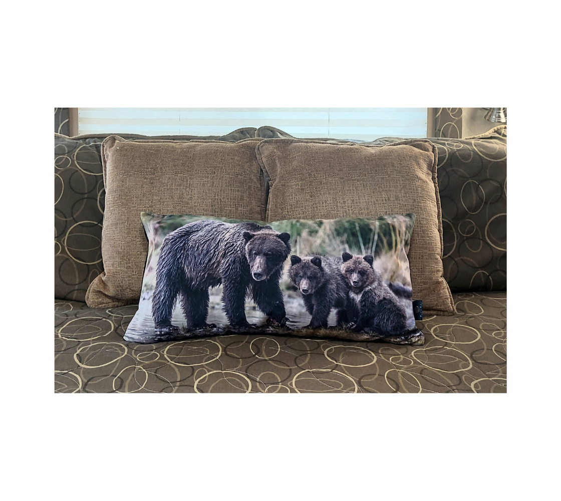 This 12 x 24 inch plush velveteen cushion cover is a photo of a dark brown grizzly mama and her 2 cubs watching us. The cushion cover background is tall green and yellow grass with the blue river's edge that the bear family is standing in. This image is of the cushion cover on a pillow form on a bed.