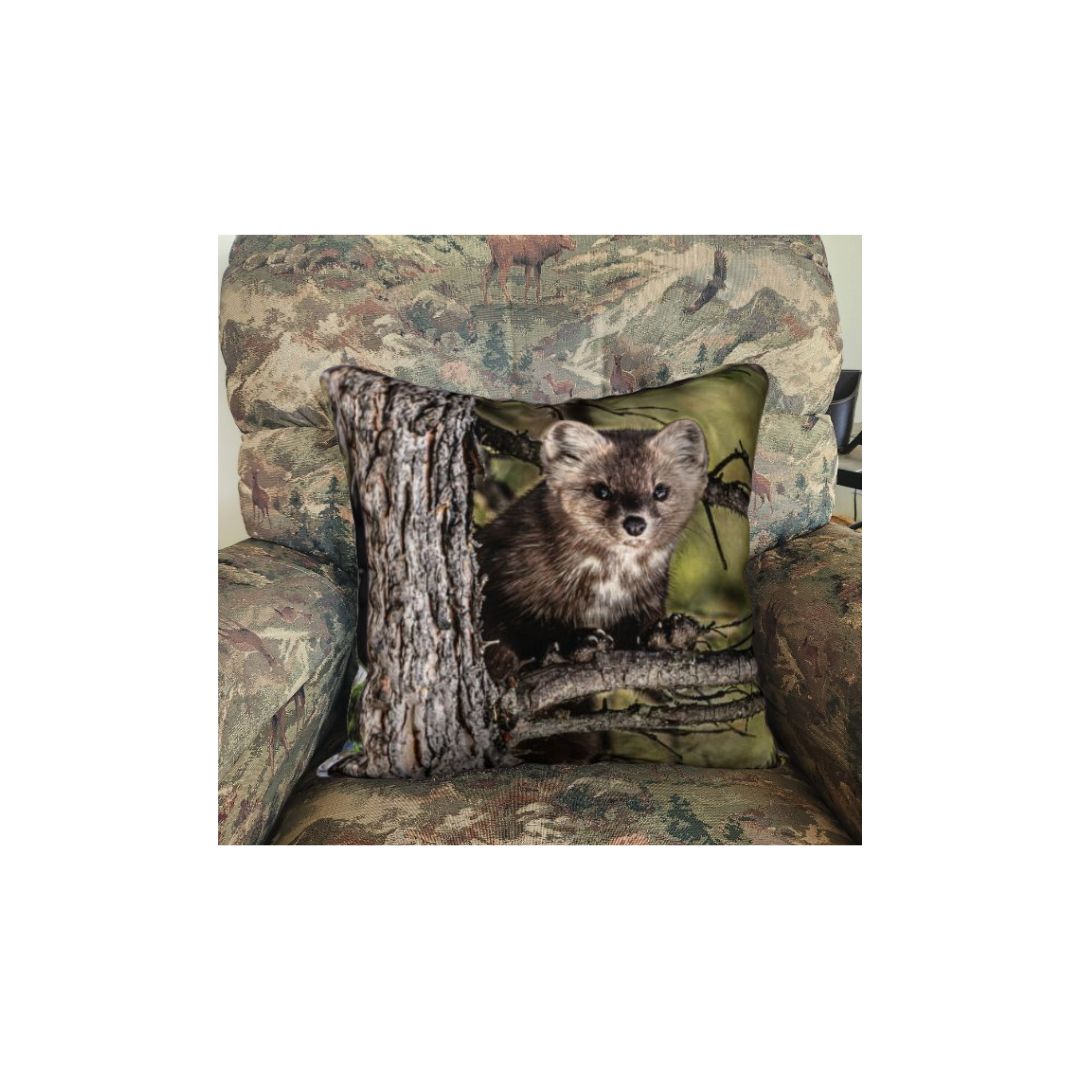 This 18 x 18 inch plush velveteen cushion cover featuring a  pine martin on a tree branch and the background is green, grey and brown. This image is of a cushion cover on a pillow form in a chair.