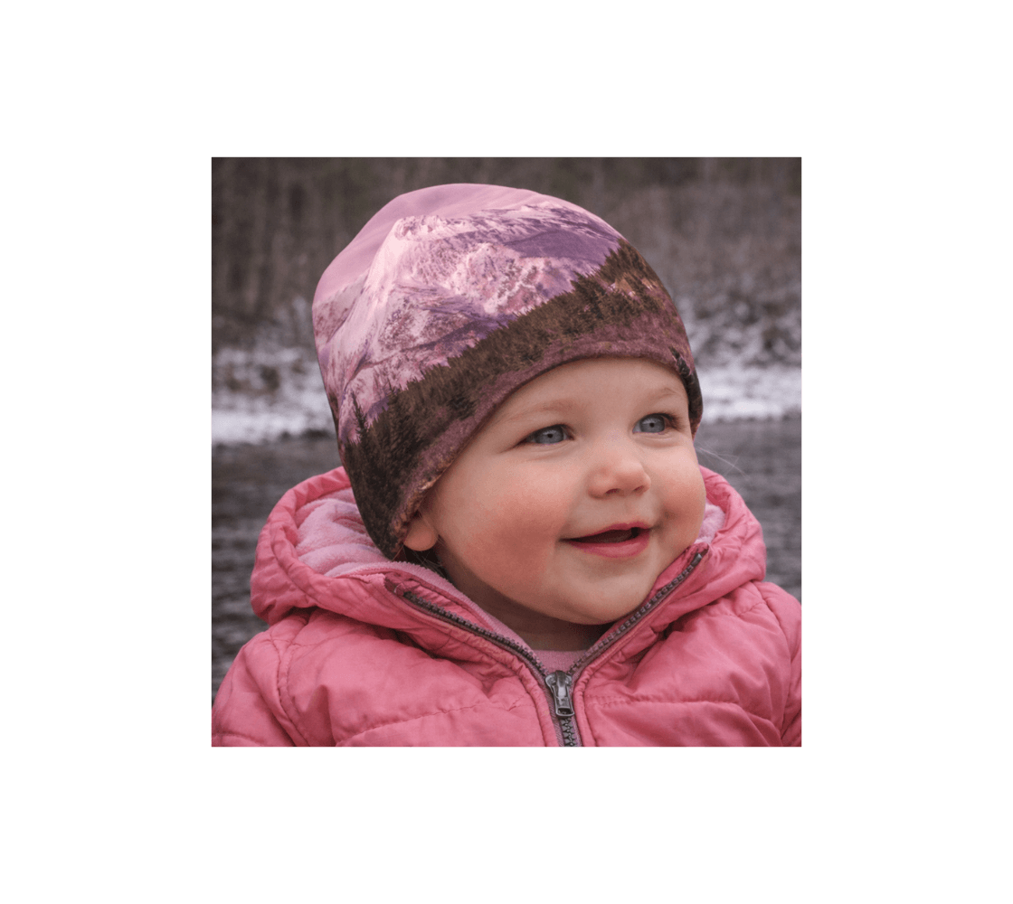 A toddler wearing a pink mountain sunset bamboo-lined beanie from our real nature photography standing by river.