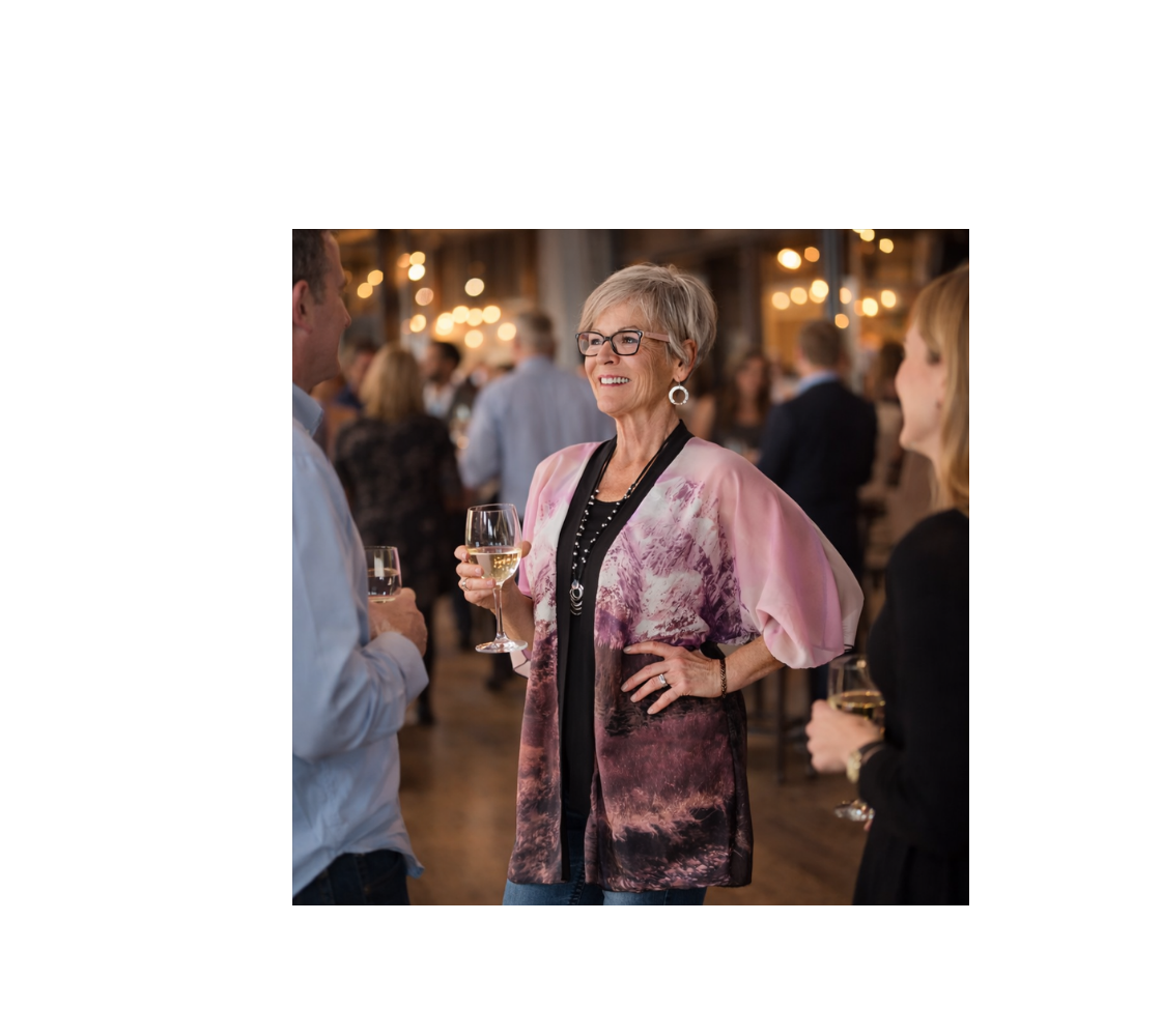 A Woman holding a glass of wine in a social setting wearing a kimono cover up with an image of a a pink mountain sunset design with a pink background. 