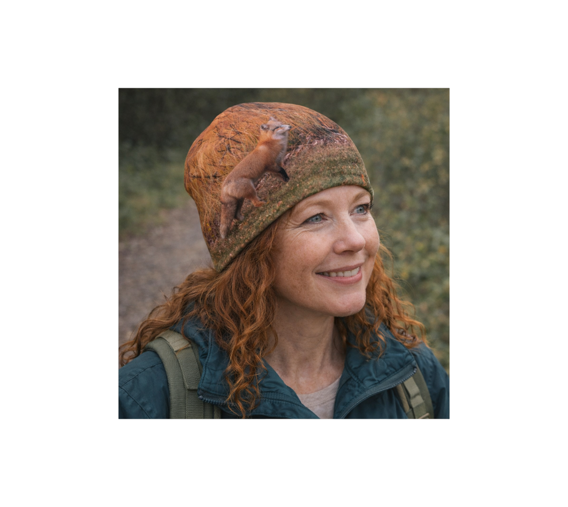 A woman with red hair standing in nature wearing a bamboo-lined beanie with an image of a red fox design with a orange background.