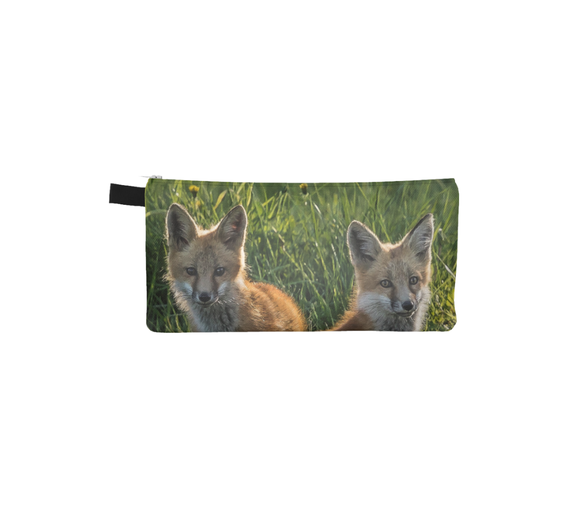 This is a photo of 2 red fox kits heads on a zippered pouch. The background is tall green grass, with the little fox kits sitting in the shadows of the nearby shade tree.