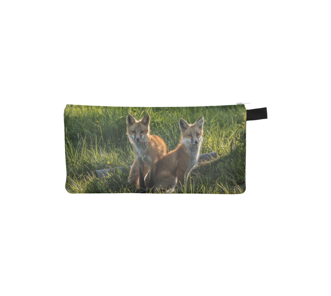 This is a photo of 2 red fox kits on a zippered pouch. The background is green grass, with the little fox kits sitting in the shadows of the nearby shade tree.