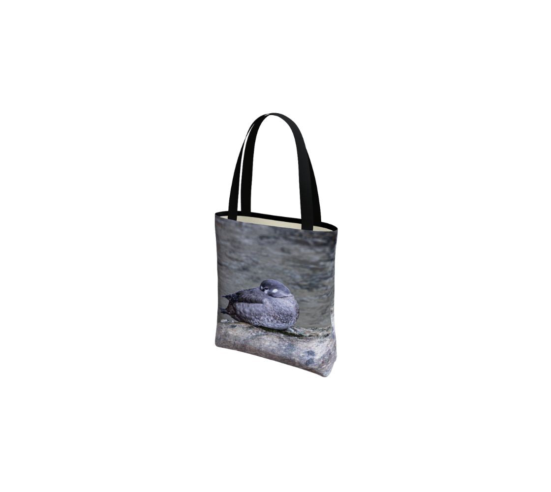 This is an image of a canvas tote bag featuring the female Harlequin Duck sleeping on a grey rock with her head tucked under its wing. The female is a more subdued brown with white patches around her eyes and on the sides of her face. The background is turbulant grey water that they prefer to be found in.