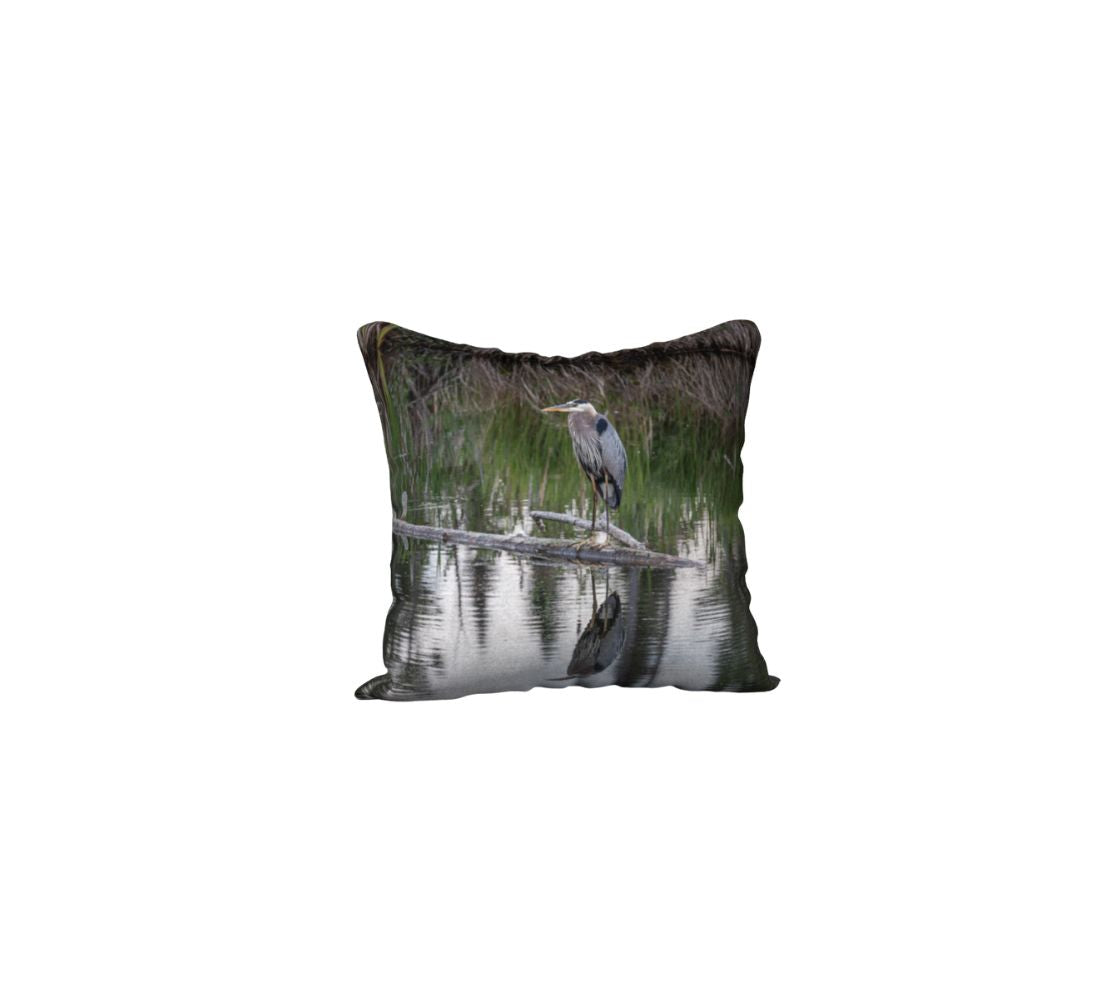 This is an image of a 18" square velveteen cushion cover featuring a great blue heron standing on a log in  pond. Standing over a meter tall, it has long blue-grey body feather, a white head, a long yellow pointed bill, and a black stripe or crest above each eye. It is standing on a grey dead branch that is lying in the blue still waters that is surrounded by tall green and brown swamp grass. The bottom of the image is a clear reflection in the water of this amazing bird.