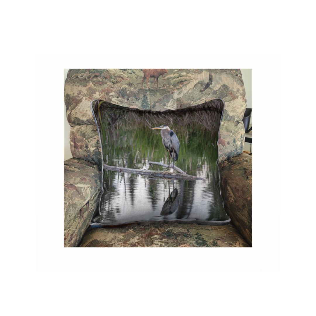 This is an image of a 18" square velveteen cushion cover in a rocking chair featuring a great blue heron standing on a log in  pond. Standing over a meter tall, it has long blue-grey body feather, a white head, a long yellow pointed bill, and a black stripe or crest above each eye. It is standing on a grey dead branch that is lying in the blue still waters that is surrounded by tall green and brown swamp grass. The bottom of the image is a clear reflection in the water of this amazing bird.