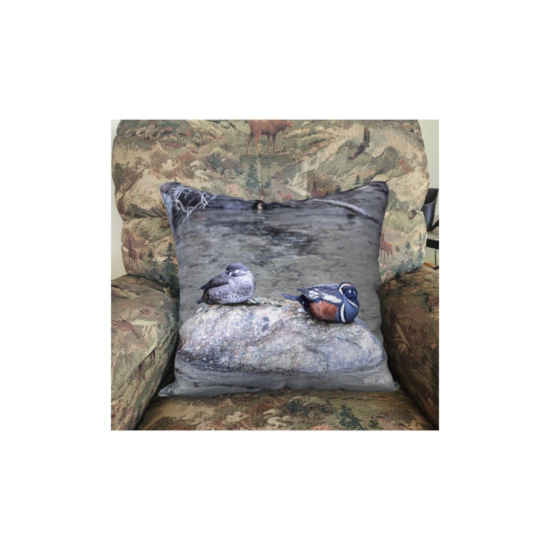This is an image of a 18" pillow in a rocking chair featuring the male and female Harlequin Ducks sleeping on a grey rock with their heads tucked under their wings. The male has a striking slate-blue plumage with chestnut flanks and distinctive white patches on its head and body. The female is a more subdued brown with white patches around her eyes and on the sides of her face. The background is turbulant grey water that they prefer to be found in.