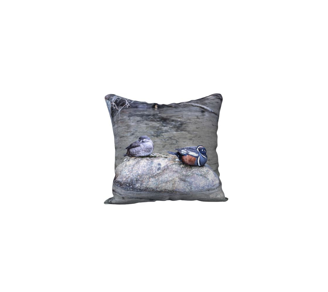 This is an image of a 18" pillow featuring the male and female Harlequin Ducks sleeping on a grey rock with their heads tucked under their wings. The male has a striking slate-blue plumage with chestnut flanks and distinctive white patches on its head and body. The female is a more subdued brown with white patches around her eyes and on the sides of her face. The background is turbulant grey water that they prefer to be found in.