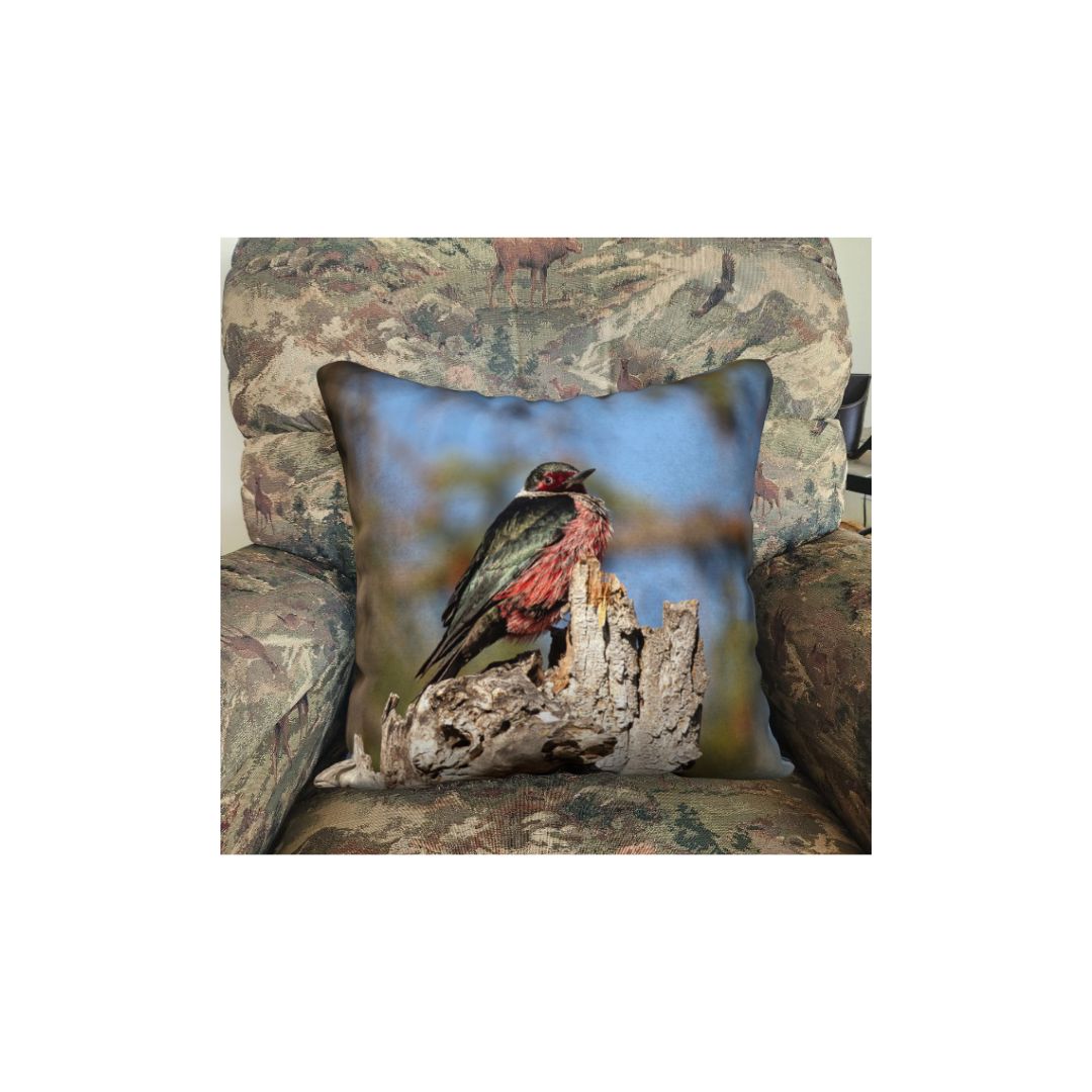 This is an image of a 18" square pillow  in a rocking chair featuring a male Lewis's Woodpecker. The bird is a medium size, with a dark green-black back, a silvery gray collar, a red face patch, and a pinkish-red belly. It is sitting a top of a broken off tree trunk that is beige in color with a background of blue sky with mottled green tree branches.