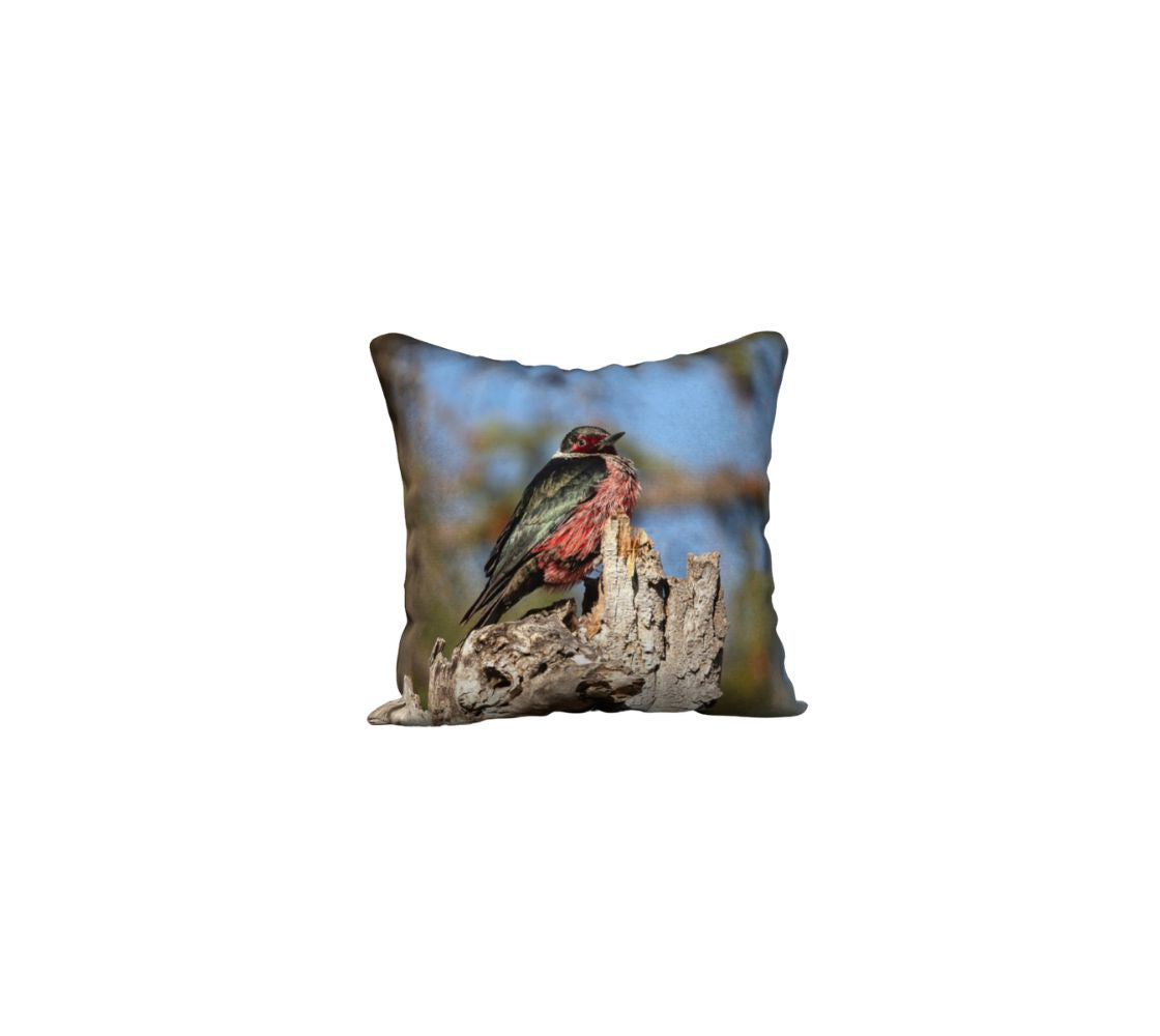 This is an image of a 18" square pillow  featuring a male Lewis's Woodpecker. The bird is a medium size, with a dark green-black back, a silvery gray collar, a red face patch, and a pinkish-red belly. It is sitting a top of a broken off tree trunk that is beige in color with a background of blue sky with mottled green tree branches.
