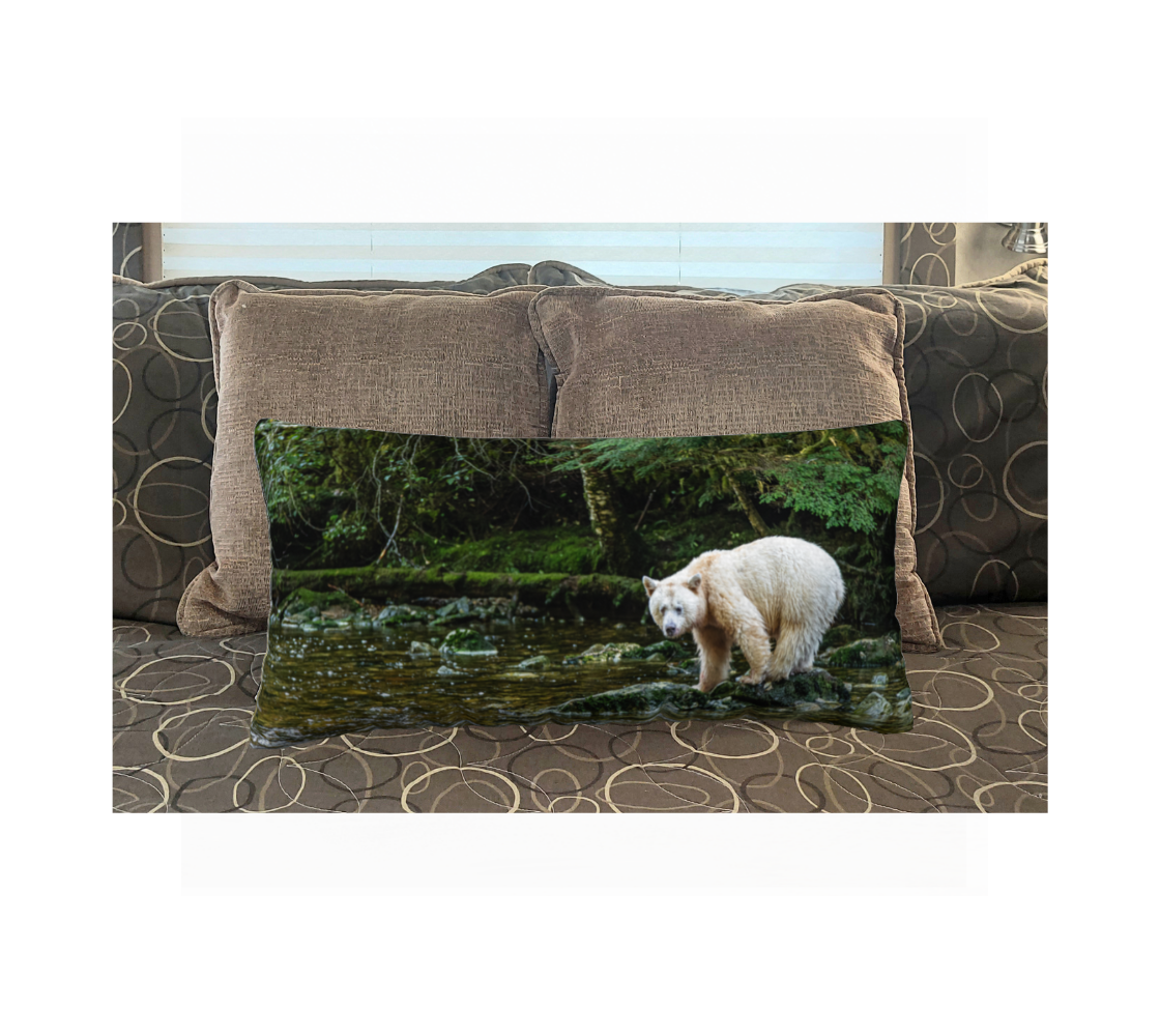 This 12 x 24 inch plush velveteen cushion cover features a white spirit bear standing on a green mossy rock watching for salmon and the background  is mossy ground cover, green tress and little bit of golden green stream. This image is of the cushion cover on a pillow form on a bed.