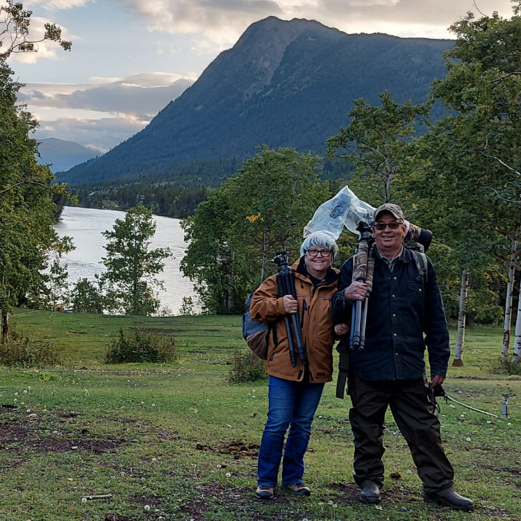 Keltie and Stan with photography gear standing in a grassy area with mountains and a lake in the background.