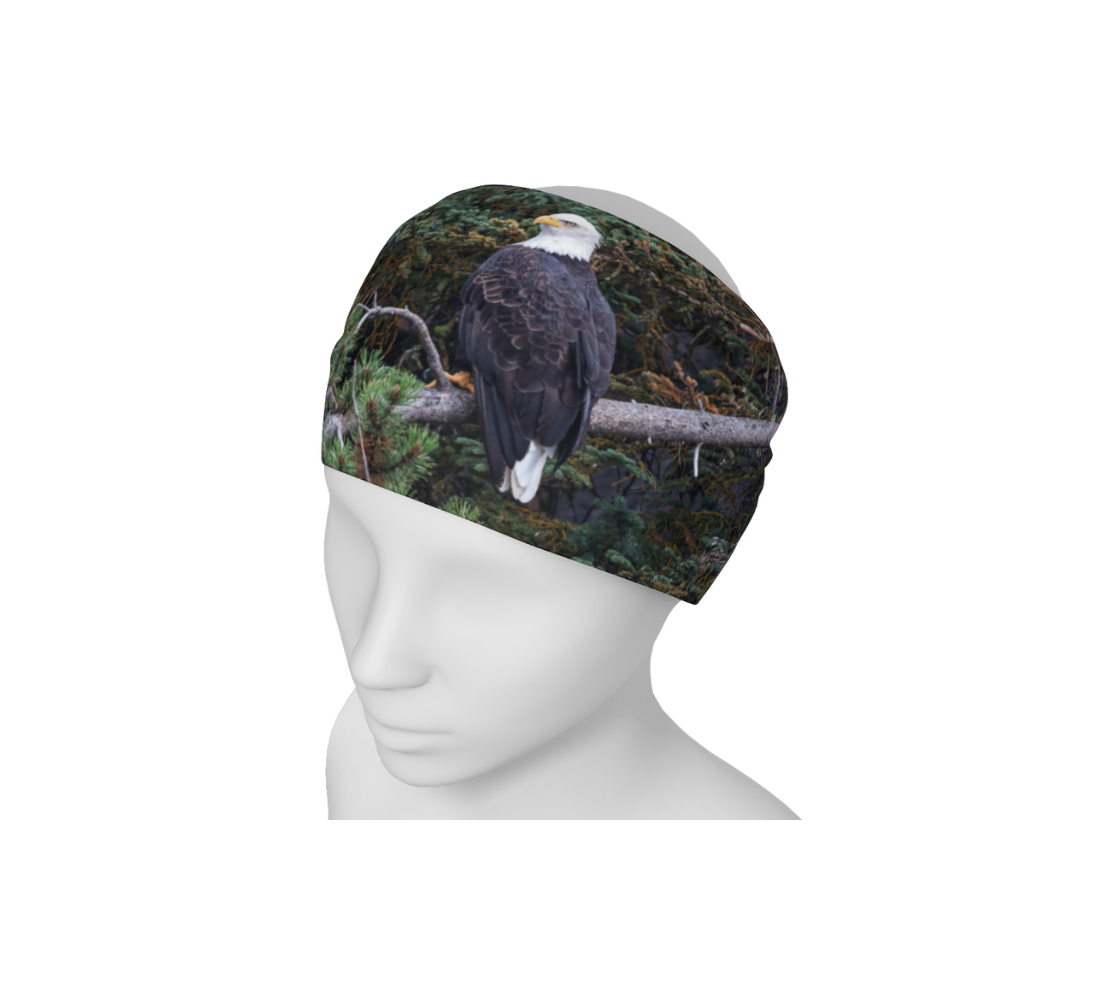 This is an image of a photo of a bald eagle on a headband on a mannequin. The background is of trees and branches in various tones of green, gold, purple and grey. The bald eagle is perched on a fallen grey tree trunk with his back to us and its head is turned slightly, as he is looking towards you. 