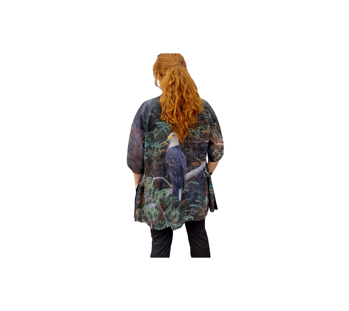 This backview is of a lady wearing a semi-sheer poly chiffon kimono robe, featuring of a photo of a bald eagle. The background is pine trees in muted colors of teal, rust and green with a bald eagle perched on a branch. Kimono has black trim and a black lightweight bamboo belt for tying.