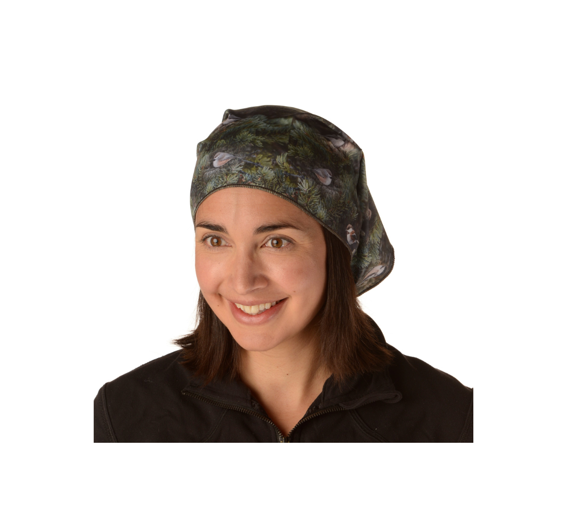 This is an image of a lady wearing a photo collage of chickadees on a head and neck band, worn as a kerchief. The fabric print is of green tree boughs in the background with a repeating collage of the four different chickadees of Western Canada in their colorful black, brown, white, rust and grey tones.