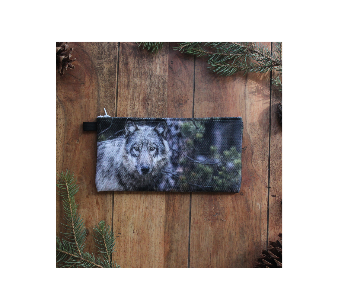 This is a durable double sided 9” x 4” canvas zippered pouch featuring real images of a grey wolf in the Rocky Mountains. On this side, the background is of evergreen trees and grey bushes, while the wolf head is full and fluffy in various tones of grey and white with beautiufl yellow eyes looking directly at you. 