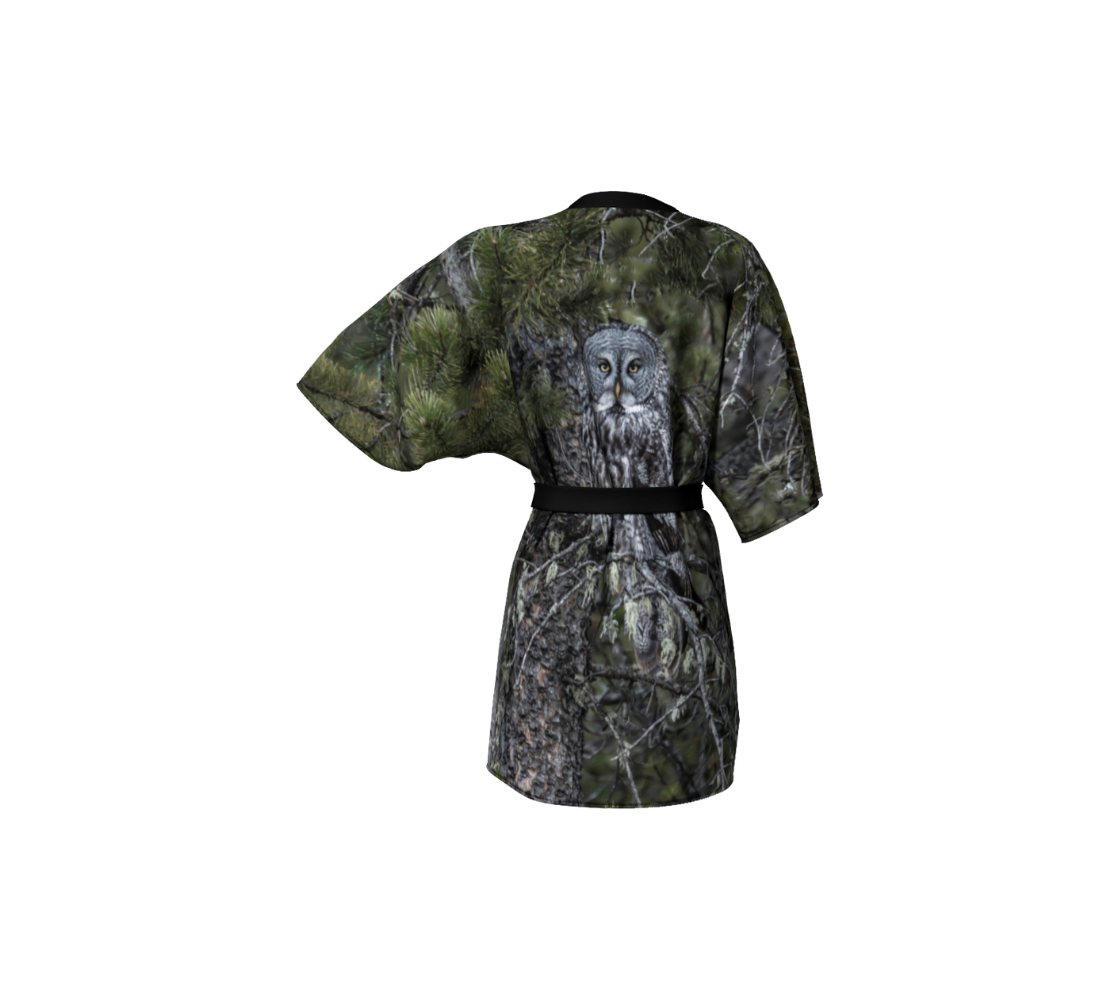 This backview is a semi-sheer poly chiffon kimono robe of a photo of an owl. The background is green pine boughs and grey pine trees, with a great grey owl perched on a branch. The  kimono has black trim and a black lightweight bamboo belt for tying.