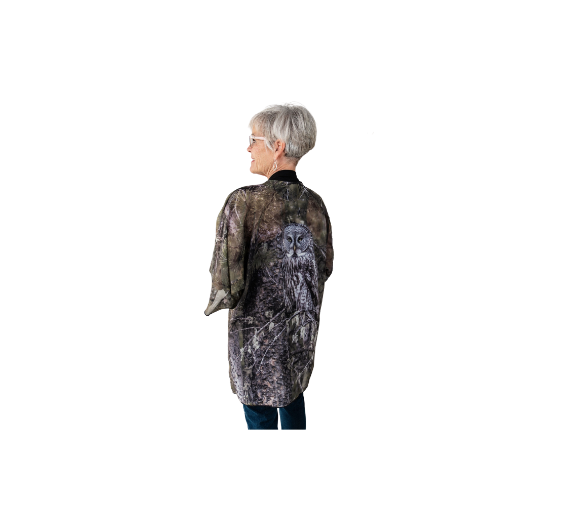 This backview is of a lady wearing a semi-sheer poly chiffon kimono robe of a photo of an owl. The background is green pine branches and grey pine trees, with a great grey owl perched on a branch. The background is green pine boughs and grey pine trees, with a great grey owl perched on a branch. The kimono has black trim and a black lightweight bamboo belt for tying.