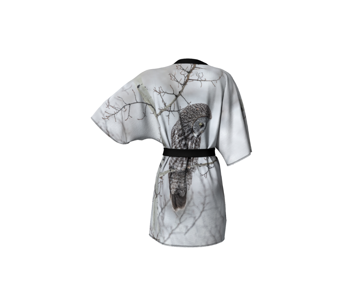 Backview of a semi-sheer poly chiffon kimono robe featuring a photo of an owl. Background is white and grey, with blurred tree branches due to the fog and frost, with a great grey owl perched on branch. Kimono has black trim with a black lightweight  bamboo belt for tying.