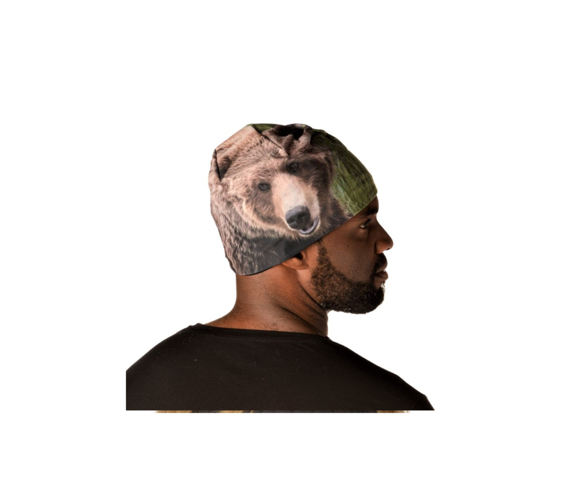 This is a side profile of a man wearing a  grizzly bear beanie. The image on the beanie is of a brown grizzly bear head, looking slightly away from you, with green grass in the background.