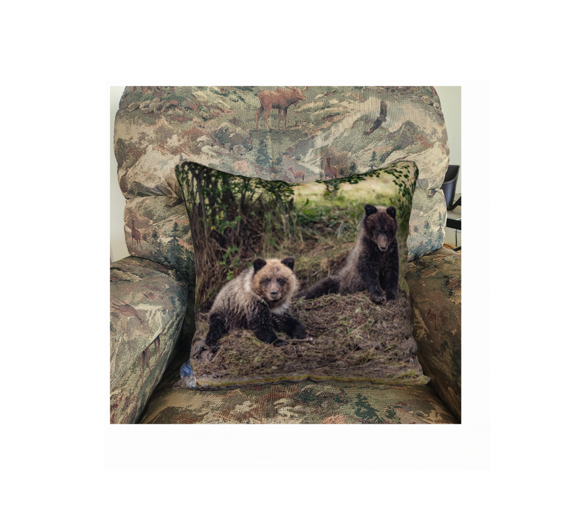 This 18 x 18 inch plush velveteen cushion cover features a photo of two dark brown and beige grizzly bear cubs on a mound of buried dead salmon. The background is green grass with yellow and brown tones of sticks and dirt. This image is of a cushion cover on a pillow form in a chair. 