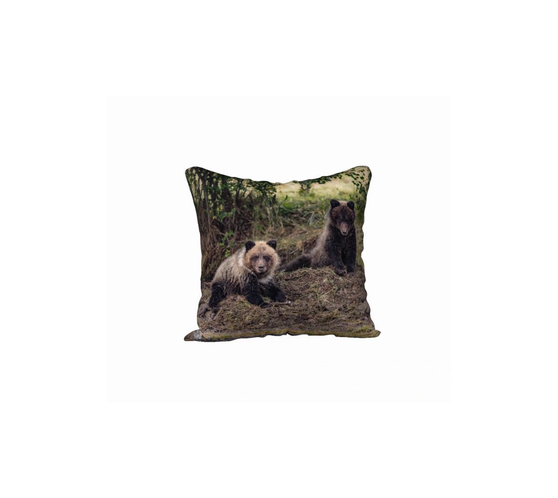 This 18 x 18 inch plush velveteen cushion cover features a photo of two dark brown and beige grizzly bear cubs on a mound of buried dead salmon. The background is green grass with yellow and brown tones of sticks and dirt.